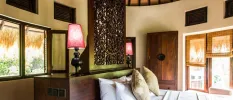 Cabana interior with an intricately carved dark-wood headboard, a wardrobe and white bedding overlooking patio doors with rattan blinds 