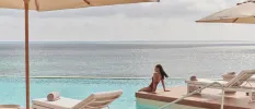 Woman in a brown swimsuit dangles her legs in a swimming pool with an infinity edge overlooking the ocean