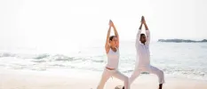 A man and woman in white active wear in a yoga pose on the beach with the ocean in the background