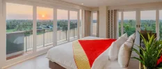 Stylish bedroom with a wraparound balcony with views of surrounding greenery and a lake, with white bedding and a red and gold throw, and a potted green plant in the corner