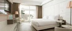 Hotel room with white bedding, wooden flooring, a neutral padded headboard and neutral lighting and cushions, with floor-to-ceiling windows overlooking a balcony