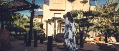 Elegant woman walking in a blue and white kaftan through a leafy courtyard towards a blush coloured stone building