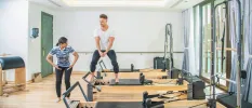 Man in white t shirt and black joggings bottoms works out on a Reformer Pilates machine as a woman next to him demonstrates
