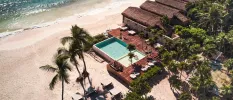 White-sand beach with turquoise shallows overlooked by a resort with thatched-roof buildings, an infinity pool and wooden deck covered in daybeds