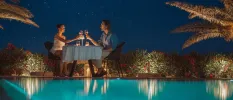 Couple toasts with wine glasses at a table set up by the poolside under a starry night sky