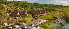 Rustic thatched-roof A-frame huts on a lakefront next to wooden decking and surrounded by jungle greenery