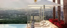 Infinity edge pool overlooking mountains and whitewashed buildings below, with sleek white-cushioned loungers and parasols topped with blue towels, and tall palm trees behind them