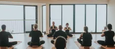 Group of people wearing black sit on blocks on yoga mats in a studio with floor-to-ceiling windows