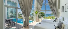 Airy room with high ceilings, white walls and bedding, window drapes and floor-to-ceiling glass overlooking a pool terrace and the ocean