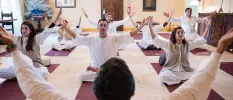 Group in loose white clothes sit cross legged with arms outstretched and eyes closed on purple yoga mats