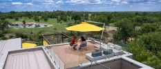 Male and female enjoy a meal under a yellow parasol on a wooden roof terrace overlooking a reserve waterhole