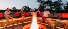 Guests relax with glasses of white wine around a central fire pit under a darkening sky