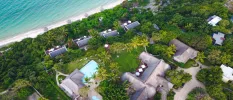 Aerial view of tropical greenery, grey-roofed buildings and a white-sand beach and ocean