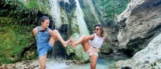 Two people touch feet while in an advanced yoga pose, with a waterfall and rocky backdrop