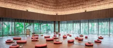 Thatched-roof pavilion with floor-to-ceiling windows, set up for a class with grey yoga mats, red cushions, blankets and bottles of water, with a gong at the head of the class