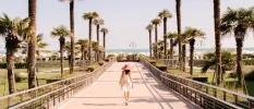 Woman in white dress and straw hat strolls across a paved bridge lined by palms towards the sea