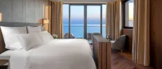 Cosy hotel room with plush white bedding, neutral curtains, a wooden floor and full-length windows looking out to sea
