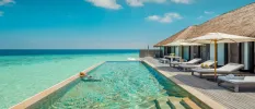 Woman relaxes in an infinity pool in a wooden deck furnished with white-cushioned loungers and umbrellas surrounded by turquoise shallows 