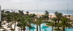 Palm trees and white parasols surrounding a swimming pool with the beach and ocean in the background