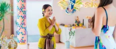 Woman in traditional dress with a flower in her hair and her hands clasped in prayer greeting a woman entering the room