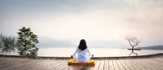 Woman sits on the edge of an infinity pool on a wooden deck meditating with misty hills in the distance