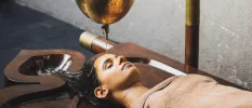 Woman lies on a dark-wood bed as an unseen therapist pours oil on her head from a gold bowl
