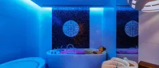Woman in black swimwear relaxing in a filled bathtub in a white room with atmospheric blue mood lighting