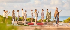 Group of men and woman in loose fitting clothes stand on a wooden platform overlooking the sea with their arms raised in a salute