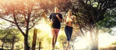 Couple in active wear jog together among pine trees as the sun rays peer through the greenery