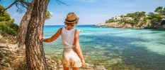 Woman in a white vest and shorts in a straw hat leans against a tree looking at turquoise waters