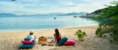 Couple gaze out to the sea as they enjoy a beach picnic on pink and blue cushions, with a blanket, picnic basket and plates of salad and fruit between them