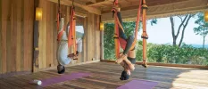 Man and woman hang from the ceiling in acrobatic ribbons, in a wooden studio space with a large window facing the sea