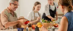 Family in aprons sit around a table spooning things out of jars, as a chef watches on