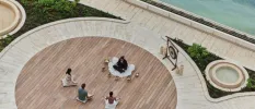 Three figures seen from above, sitting cross legged on a round wooden deck as an instructor surrounded by sound bowls and with a gong behind her, takes a healing class
