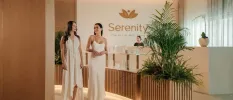 Smiling women in white dresses stroll through a reception area with wood-panelled walls, soft lighting and plant beds, with a sign that reads 'Serenity the art of wellbeing' behind them