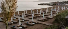 Sandy beach with wooden walkways, cushioned loungers and white parasols, looking out onto a curved bay under a cloudy sky