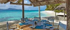 Thick wooden table set up with blue napkins and place mats under a thatched roof overlooking the ocean