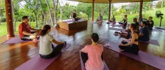 Group sit cross legged on mats in a circle in a open-sided yoga pavilion, as an instructor demonstrates on a raised block upfront 