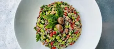 White bowl with grains, tomatoes, edamame beans and cucumber, with a mint and olive garnish