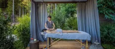 Woman lies partially covered by a blue towel on a massage bed in an open-air cabana surrounded by a forest, as a therapist massages her back