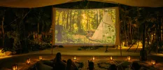 Group sit on blankets and cushions on the floor with lanterns all around as a film plays on a giant screen