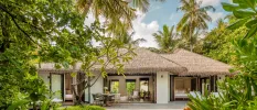 Thatched-roof villa surrounded by palm trees and shrubs, with a wooden deck furnished with a table and chairs