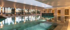 Indoor swimming pool with soft lighting, white loungers lined up one one side and full-length windows