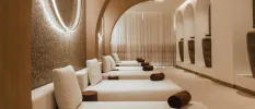 Softly lit relaxation room with a line of white-cushioned beds, a tiled wall, archways decked out with oversized vases and brown rolled up towels on each bed