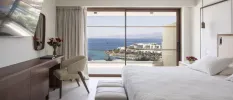 Minimalist hotel room with crisp white bedding, walls and floors, a dark-wood dressing table and wall mirror, a wall-hung TV and floor-to-ceiling windows overlooking a balcony with a sea view