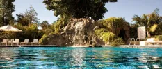 Huge pool with rockery and a waterfall in the middle, surrounded by tropical gardens, white parasols and loungers