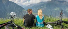 Man and woman in active wear take a break from cycling sitting down on a hillside overlooking the mountains