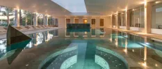 Whirlpool section in an indoor pool, surrounded by white loungers and full-length windows with gardens one side, and seating areas the other