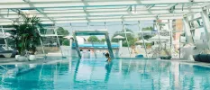 Woman in a black swimsuit relax in an indoor pool that leads outdoors with wraparound glass windows and ceiling