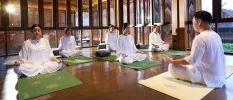 Group in white on green mats with closed eyes, legs crosses and their palms upwards, in a wood-walled, wooden floored studio